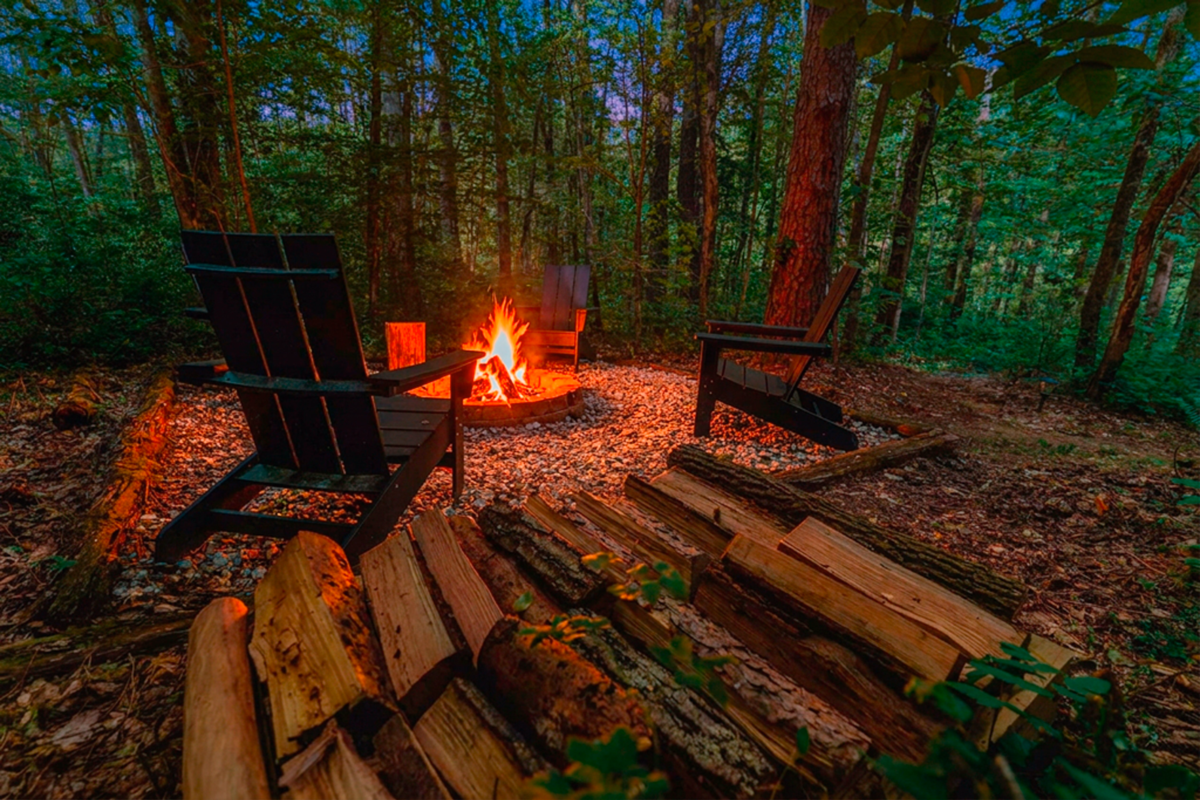 A glowing woodland fire pit surrounded by Adirondack chairs at twilight